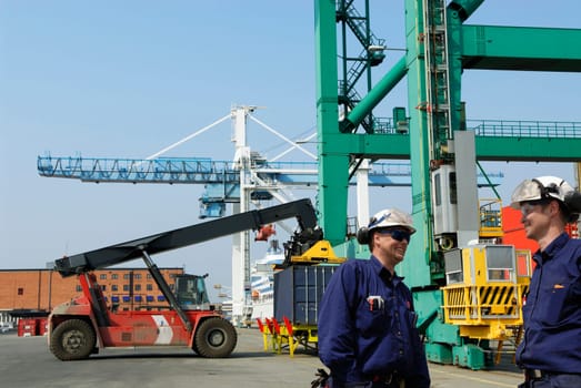 busy container-port, trucks stacking containers from crane, two port-workers in foreground