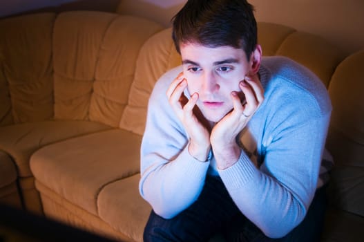 Head and shoulders shot of a young man with hands on his cheeks looking at the TV in the darkened living room