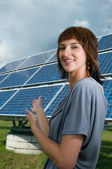 Upper body shot of a young brunette woman in casual casual dress on a green meadow in summer pointing to a photovoltaic system behind her