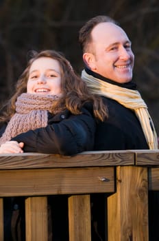 Upper body exterior shot of father with teenage daughter back to back on railing of wooden bridge smiling at camera