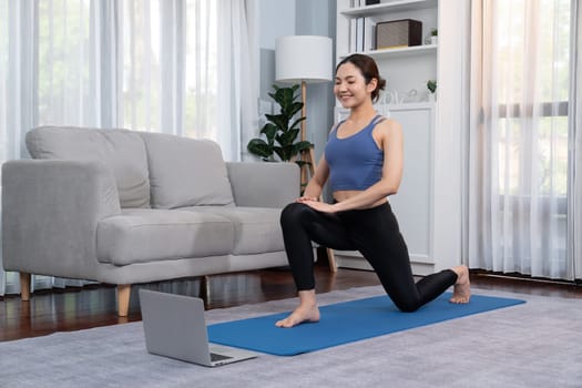 Asian woman in sportswear doing yoga exercise on fitness mat as her home workout training routine. Healthy body care lifestyle woman watching online yoga video on laptop. Vigorous