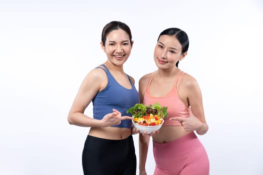 Two young sporty Asian women in sportswear holding salad bowl fill with fruit and vegetable. Natural youthful and fit body lifestyle people with balance nutrition on isolated background. Vigorous