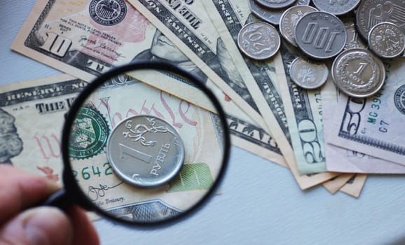 A hand holds a magnifying glass over one metal Russian ruble against the background of dollar bills. Default in Russia and strengthening of ruble.