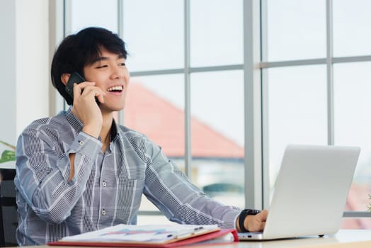 Asian businessman smiling and talking on a mobile phone at home office desk. The man smile call on smartphone with customer at the house, making sell offer