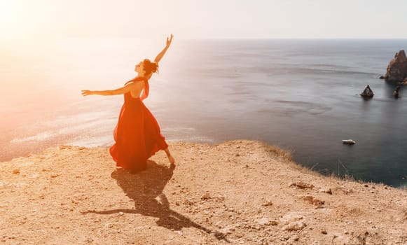 Side view a Young beautiful sensual woman in a red long dress posing on a rock high above the sea during sunrise. Girl on the nature on blue sky background. Fashion photo.