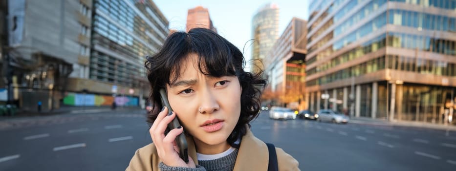 Close up portrait of concerned asian woman, talks on mobile phone and hears bad news, looks worried, feels frustrated by telephone conversation, stands on street.