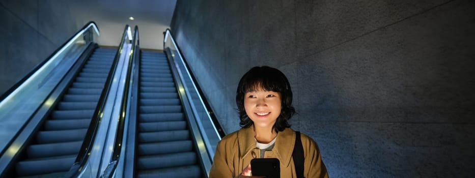 Portrait of happy beautiful asian girl, standing with smartphone on escalator, going down to metro station, commuting via tube.