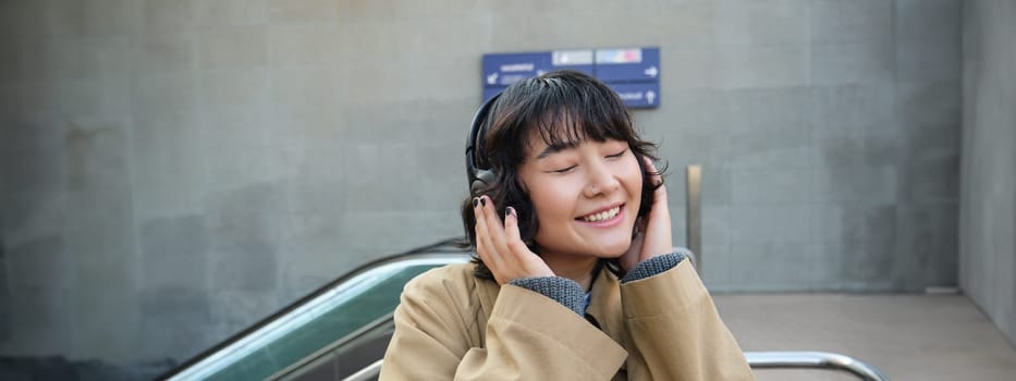 Portrait of happy woman, korean girl in headphones, listening music in headphones, enjoying sound of earphones, laughing and smiling.