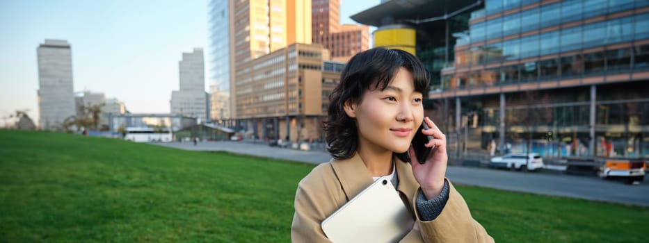 Stylish smiling girl, university student in trench coat, holds tablet, talks on mobile phone, has conversation over telephone and looks relaxed, stands on street.