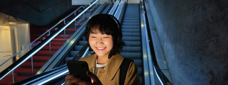 Portrait of girl student in headphones, listens music, commutes, goes down escalator, looks at smartphone with pleased smile, happy face.
