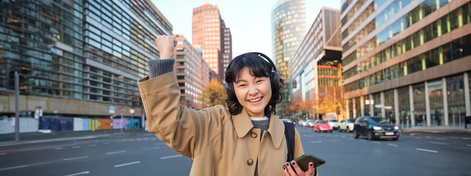 Young happy woman celebrating on street, holding smartphone and cheering, reacts amazed to good news, posing happy in city centre.