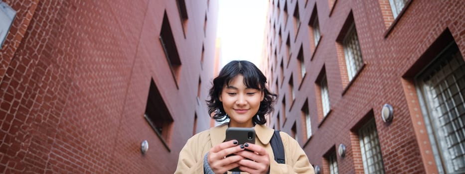 Lower angle view of brunette korean girl, listening music in headphones, walking along street and looking at smartphone, reading message on mobile phone.