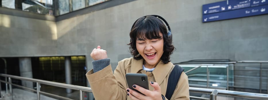 Happy asian girl in headphones, looking at mobile phone and celebrating, reading message with good news, screams yes with excitement, stands on street.