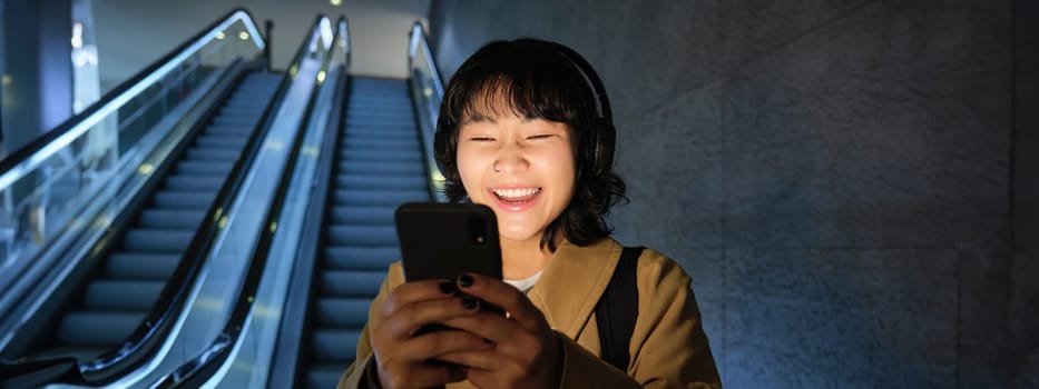 Laughing cute Korean girl on escalator, going down to tube, underground or metro, using mobile phone, watching smth funny on smartphone app.