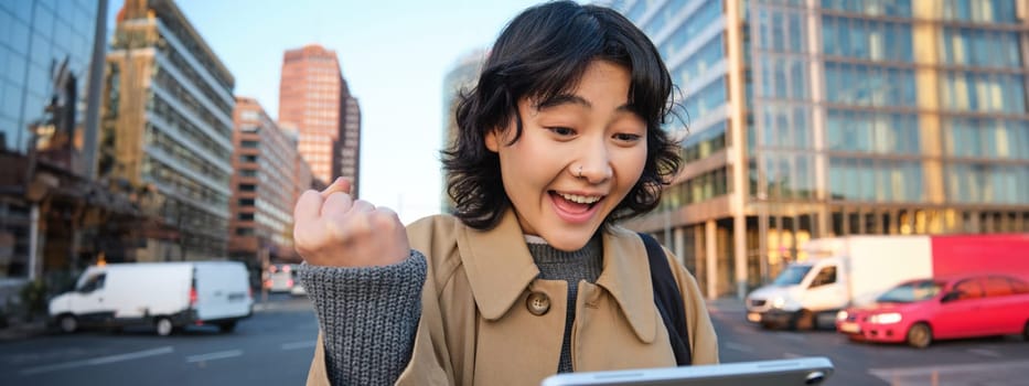 Enthusiastic asian girl with digital tablet, smiling and looking amazed, happy with something good, holding digital tablet, standing on street.