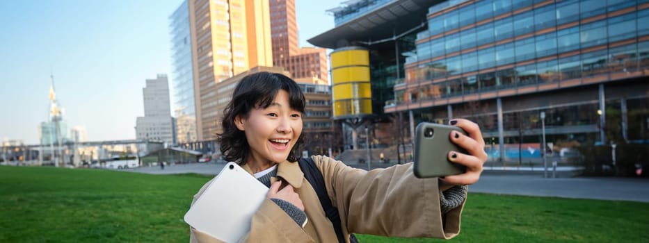 Happy young woman, student takes selfie on street, holds her tablet and homework notes, uses smartphone. Copy space