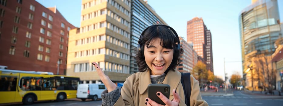 Young happy woman celebrating on street, holding smartphone and cheering, reacts amazed to good news, reads phone text message with surprised joyful face.