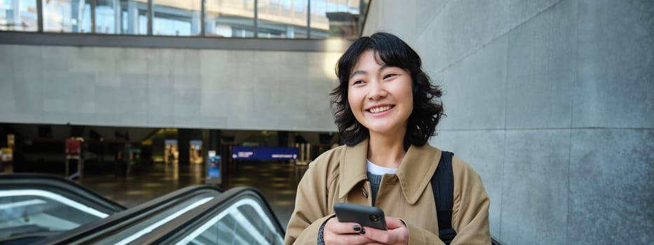 Portrait of cute korean girl in trench coat, going up escalator, holds mobile phone and smiles pleased, commutes.
