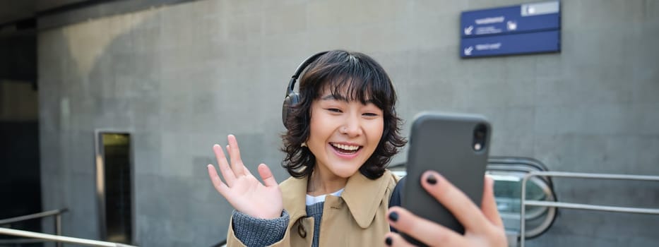 Stylish hipster girl in headphones, says hello to mobile phone camera, waves hand at smartphone, video chats, records a blog, listens music while stands on street.