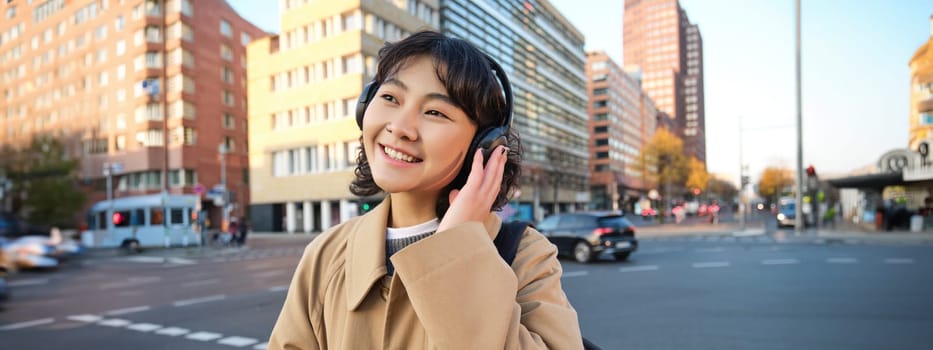 Portrait of young smiling korean girl, walking along city centre, listening music in headphones and holding mobile phone. Copy space