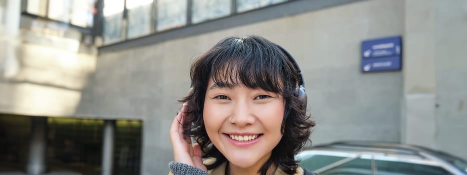 Close up of smiling brunette girl in headphones, listens music, travels around city, commutes to work, stands on a street.
