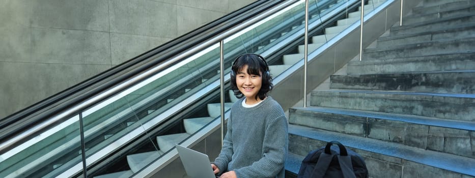 Portrait of beautiful young korean woman, student sits on stairs in public place, listens music in headphones, works on project on laptop, works remotely.