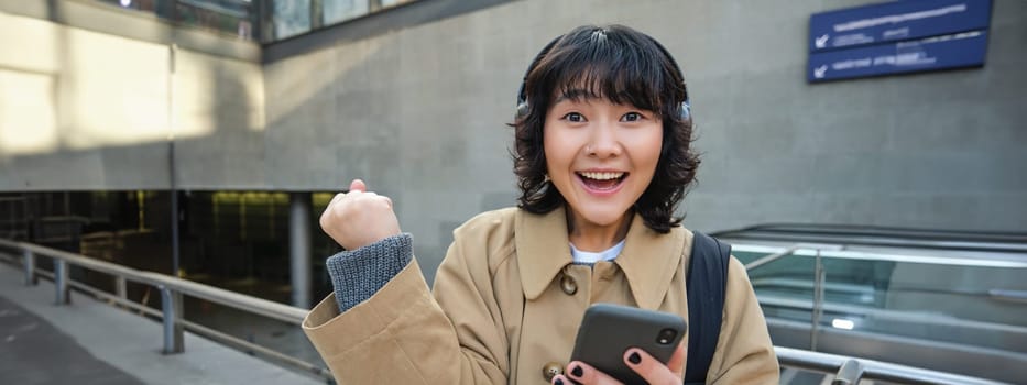 Beautiful brunette girl in trench coat, listens music in headphones and holds mobile phone, waits for someone on street and drinks coffee.