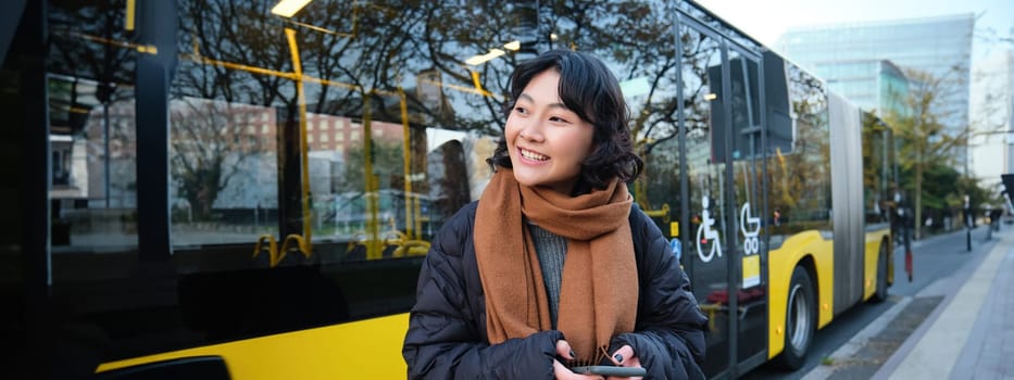 Portrait of korean girl buying ticket for public transport online, using mobile application on bus stop, wearing winter clothes.