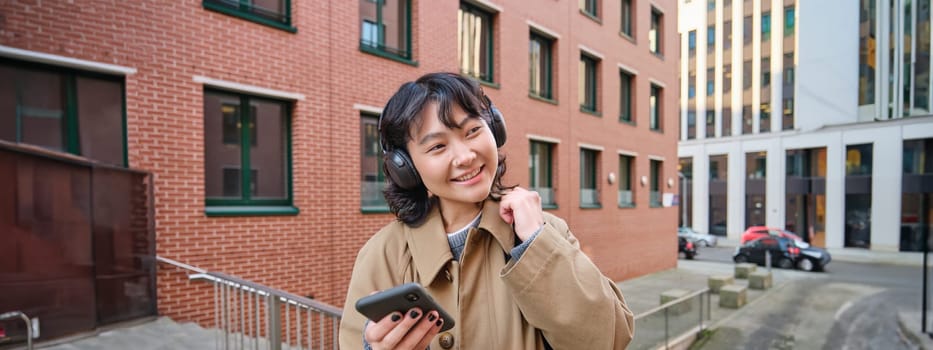 Young happy woman in trench coat, listens music in headphones, holds smartphone, uses mobile application to find route in new city, stands on street with backpack.