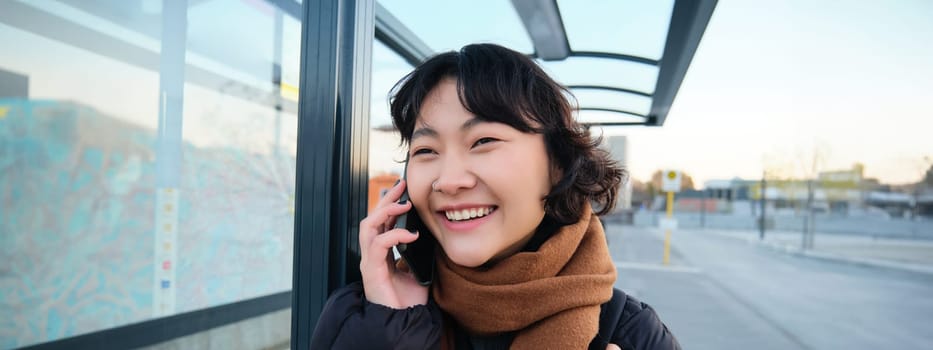 Close up of cute Korean woman, making a phone call, talking and smiling on telephone, standing in winter jacket on bus stop, waiting for her transport to arrive.