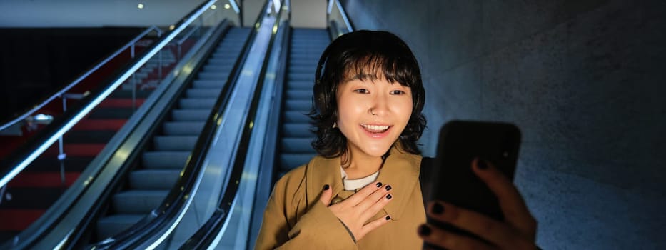 Portrait of asian girl uses smartphone, listens music in headphones, goes down escalator in city centre in evening, looking surprised at phone, gasping amazed.