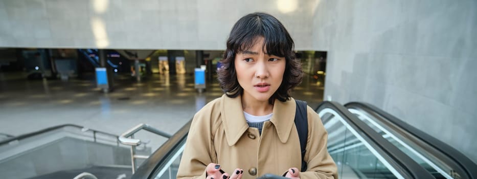 People in city. Portrait of korean girl standing on escalator, looks confused after reading text message on mobile phone.