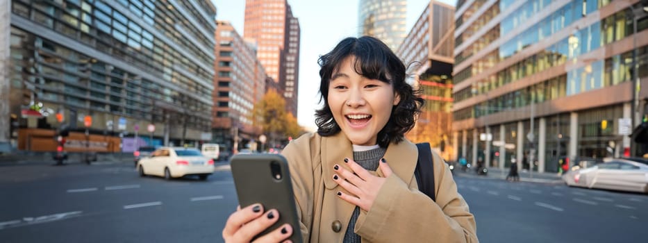 Image of korean girl video chats with smartphone, looks at her phone with surprised and amazed face, hears great news, stands on street.