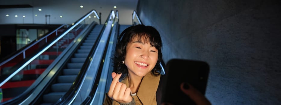Stylish smiling asian girl, taking selfie on mobile phone while riding escalator, going down to metro station, showing finger heart and posing for photo.