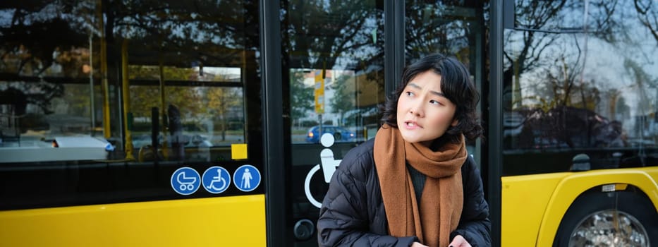 Korean girl in winter clothes, waits for her bus on stop, looking for her transport, holding mobile phone, checking timetable on smartphone application.