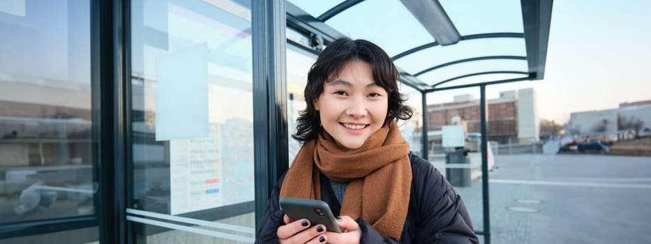 Cute young girl, student waiting on a bus stop, looking for her public transport on app tracker, holding smartphone, standing on street near road.