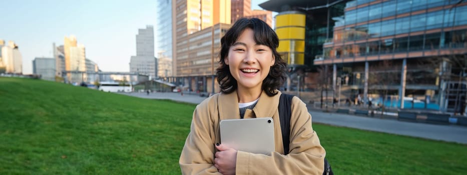 Image of korean girl with happy face, walks around town with student tablet, stands on street and smiles. Copy space