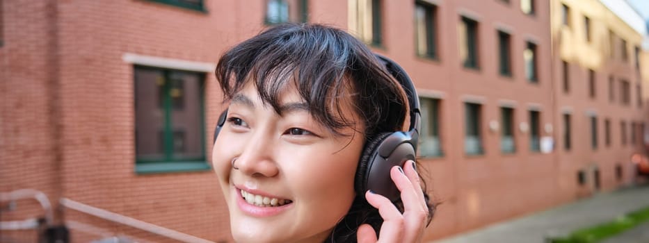 Close up portrait of stylish brunette asian girl, listens music in headphones, touches earphones and smiles, enjoys favorite song, stands on street.
