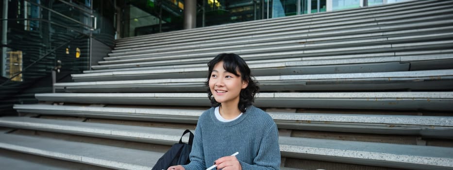 Smiling girl, graphic designer, using digital tablet and pen tool to draw, does home assignment for university, sits on stairs.