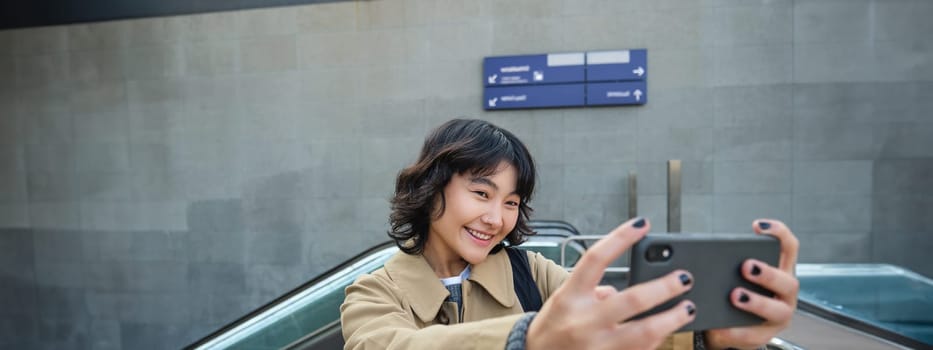 Portrait of stylish korean girl, student takes selfie on street, holds smartphone and poses on its camera, makes video on tourist trip, smiles with joy.