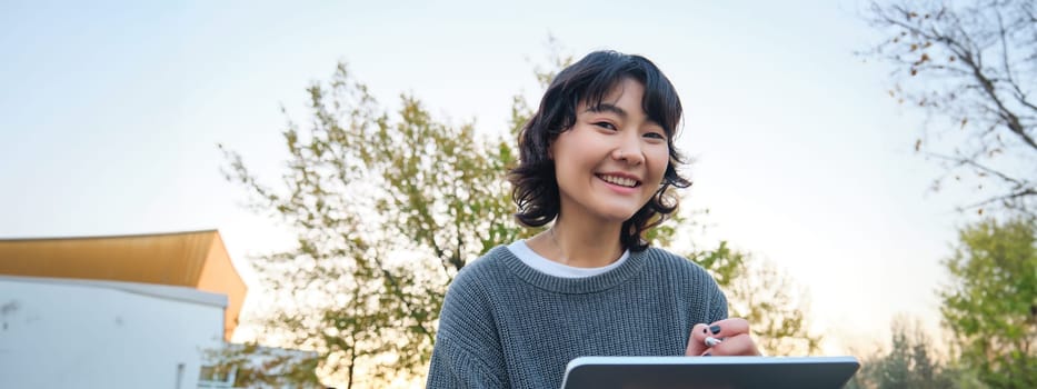 Close up portrait of young korean girl sits outdoors in park, holds her digital tablet and graphic pen, draws scatches, gets inspiration from nature for art, smiles happily.