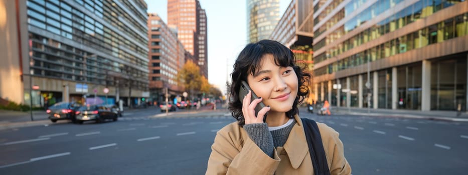 Smiling asian girl makes a phone call, stands on an empty street, calling someone on telephone, waiting for friend in city, going to a meeting.