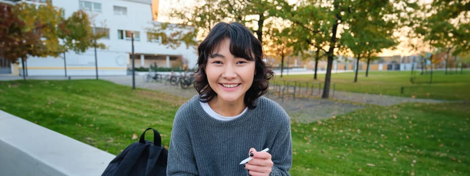 Young asian girl with graphic pencil and tablet, sits in park on bench, draws scatches, does her homework outdoors.