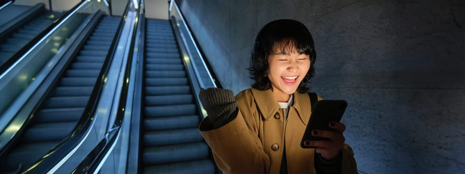 Urban lifestyle and people. Portrait of girl feeling happy and excited, going down escalator in headphones, looking at smartphone and celebrating.