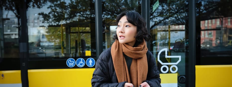 Portrait of korean girl looking for her bus on a stop, holding mobile phone, checking schedule, time table on smartphone app.