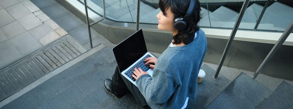 Close up portrait of girl, student works on laptop and listens music in headphones. Blank computer screen and hands typing on keyboard.