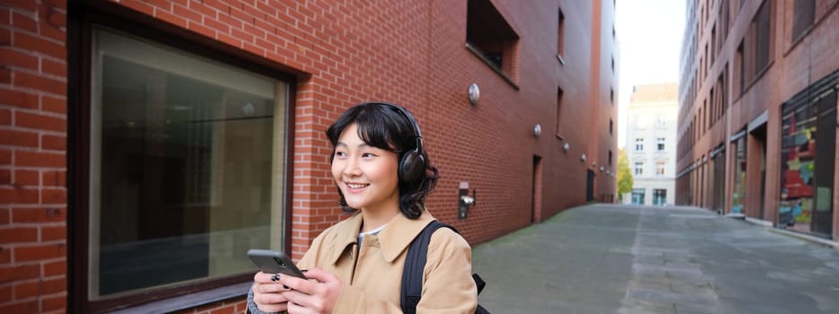 Beautiful smiling woman walking around town, listening music and using mobile phone app, going to college, wearing headphones.