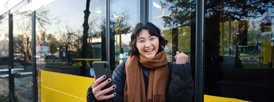 Enthusiastic asian woman, standing on bus stop with smartphone, looking at phone screen with amazed, triumphing face, winning, hear great news on video chat.