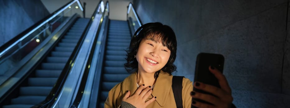 Stylish hipster girl in headphones, going down escalator and video chats, records herself on smartphone, looks at mobile phone camera.