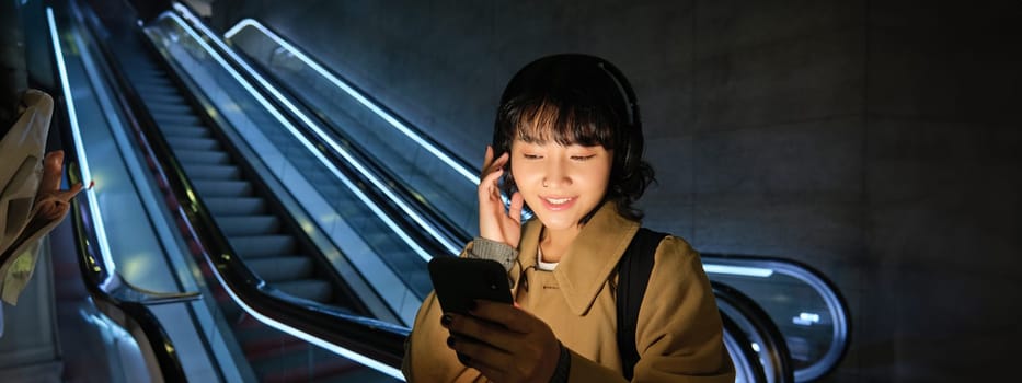 Young girl in headphones looks at her mobile phone, stands near escalator, listens to music while travels in city.